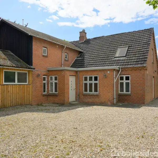 Et rødt murstenshus står stille omgivet af en gårdsplads med grus. Huset har et mørkegråt tegltag, og en blå himmel er synlig i baggrunden.