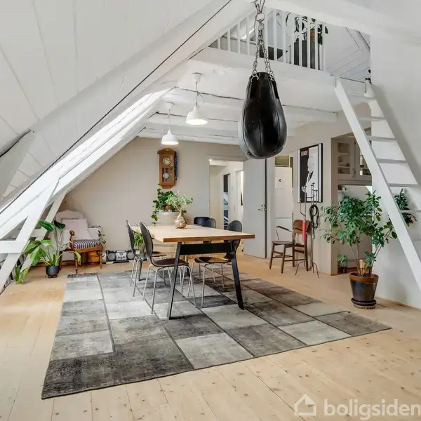 En spiseplads med et træbord og sorte stole står under et skråt loft. En boksebold hænger fra loftet og en stige fører til en hems. Rummet er lyst med grønne planter.