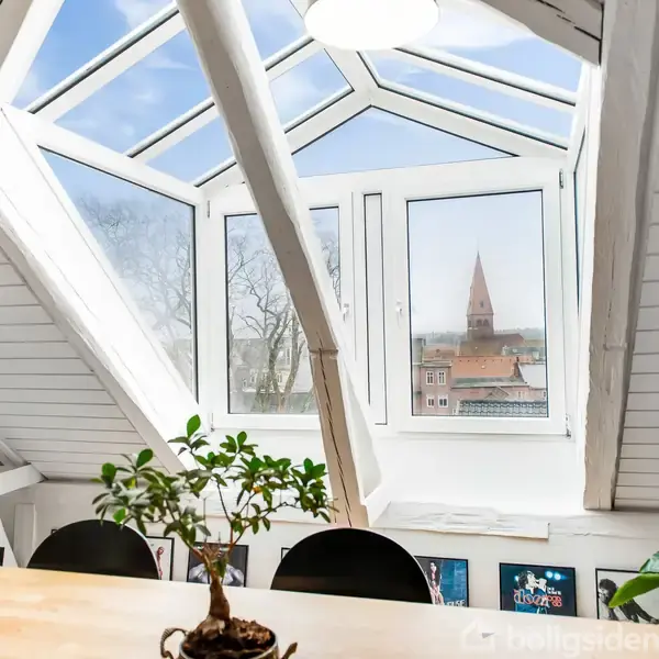 Et loftsværelse med stor tagvindue åbner mod en blå himmel og byudsigt med en kirkespir. Et bord med en lille plante og stoler står i forgrunden.