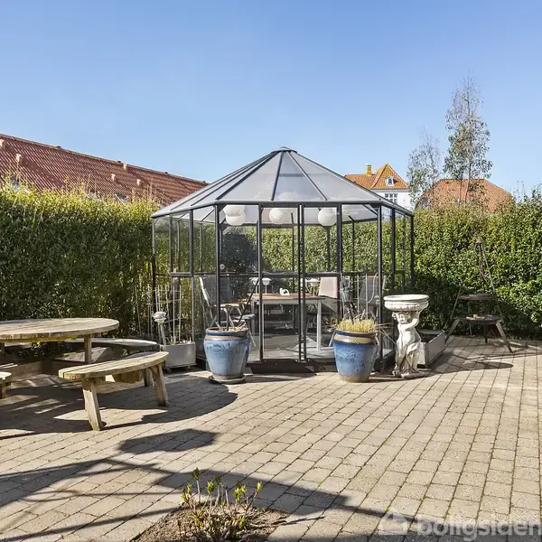 Rundt havebord med bænke står på flisebelagt terrasse. Drivhus i baggrunden omgivet af hække. Flere krukker med planter og fuglebad ved siden af. Klare, blå himmel over haveredskabsrum.