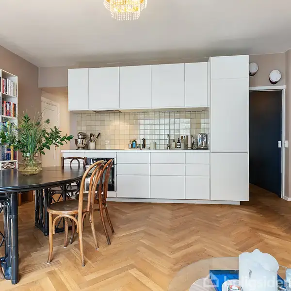 Et moderne køkken med hvide skabe og fliser. Et rundt spisebord med træstole står forrest. Der er en stor reol med bøger til venstre og en sofa til højre.