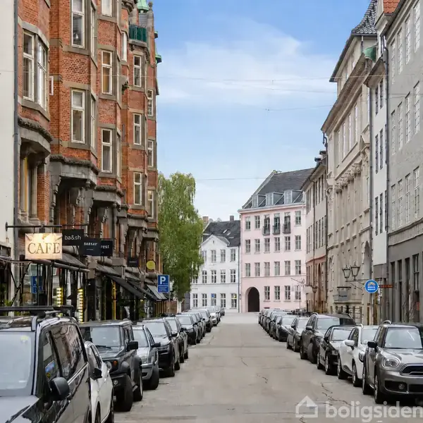 Gade fyldt med parkerede biler på begge sider, flankeret af historiske bygninger. Et skilt på en bygning til venstre viser teksten "CAFE". Baggrunden viser en hvid, klassisk bygning.