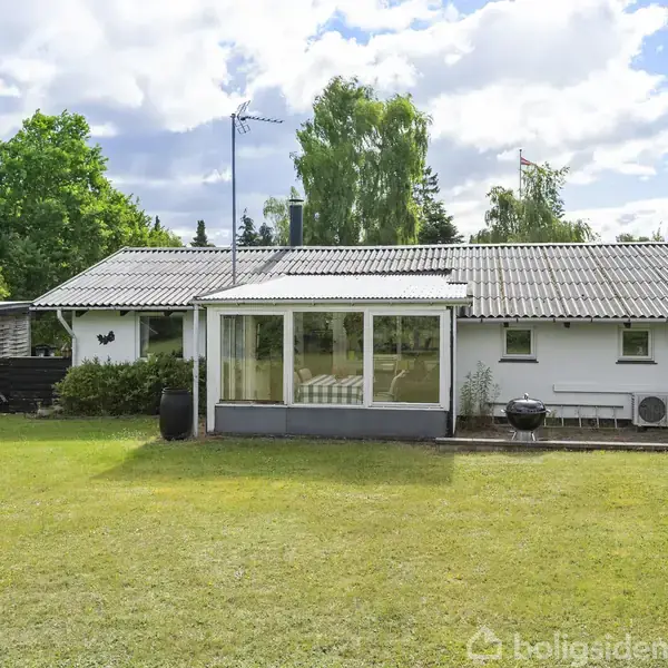 Et hvidt hus med en veranda står på en græsplæne. Solen skinner, omgivet af træer og buske i en have. Skyer ses på himlen. Ingen tekst er til stede.