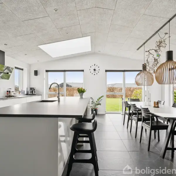 Et moderne køkken-alrum med en stor køkkenø og integrerede apparater. På højre side er der et spisebord med sorte stole. Store vinduer giver udsigt til en solrig have.
