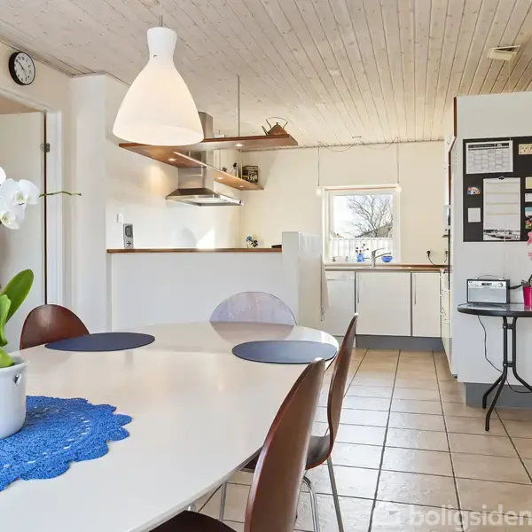 Et spisebord med hvide orkidéer på en blå dug står centralt. Rummet er lyst med trægulv og -loft, et åbent køkken i baggrunden og en sort tavle på væggen.