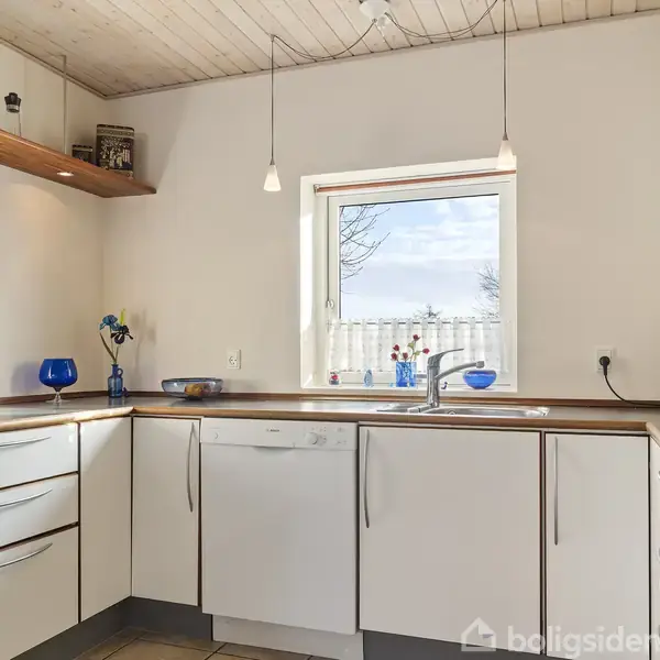 Et lyst køkken med hvide skabe og træbordplader. Vinduet centralt, udsigt til udendørs hegn. Loftshylde med krukker, to hængelamper over disken, flisegulv. Ingen tekst.