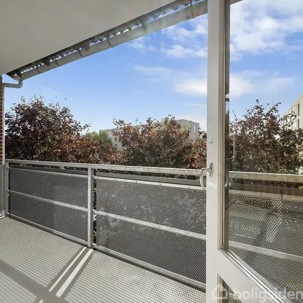 Altanen er omgivet af metalgelænder og vender mod træer med rødlige blade, under en blå, let skyet himmel. Bygninger er synlige i baggrunden.