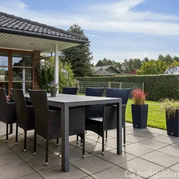 En sort spisebordsstol og bord står stationært på en flisebelagt terrasse ved et hus. I baggrunden ses en græsplæne og buske i en haveindstilling under en blå himmel.