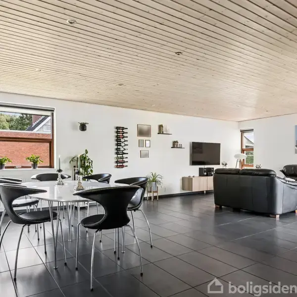 En rummelig stue med sorte fliser, hvor et spisebord med sorte stole står til venstre. En sort sofa og et tv er placeret til højre. Væggene har billeder og planter.