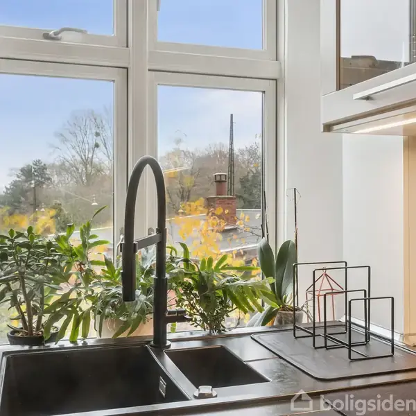 En sort køkkenvask med høj tud og planter i vindueskarmen. Køkkenredskaber hænger på væggen, og udenfor ses et grønt landskab gennem vinduet.