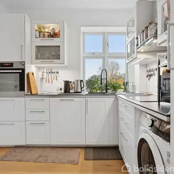 Et moderne køkken med hvide skabe og apparater som ovn og vaskemaskine. Der er en kedel og planter ved vinduet, omgivet af lyse vægge og trægulv.