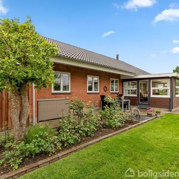 Rødt murstenshus med fladt tag, har en velholdt have med blomsterbede og græsplæne omkring. Foran huset står et stort træ, og i baggrunden er der blå himmel med få skyer.