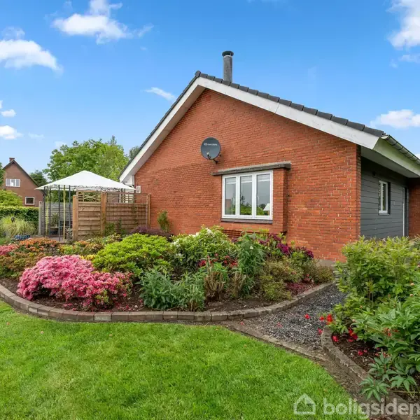 Rødt murstenshus med vindue og parabol. Omgives af velplejet have med blomsterbede, buske og græsplæne. Blå himmel med få skyer i baggrunden.