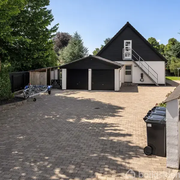 En sort-og-hvid bygning med en trappe udenfor, beliggende i en gårdsplads med brosten. To affaldscontainere står tæt på, omgivet af træer og en hæk.