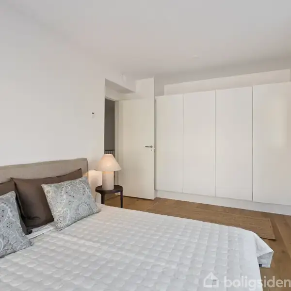 Soveværelse indeholder en seng med hvidt quiltet sengetæppe og mønstrede puder. En lampe står på sengebordet. Værelse har en stor garderobe og en vægspejl i en minimalistisk indretning.