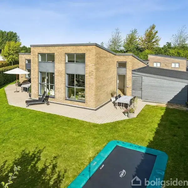 Et moderne murstenshus med store vinduer står i en velplejet have. Der er en terrasse med møbler og en nedgravet trampolin. Træer omkranser ejendommen under en klar blå himmel.