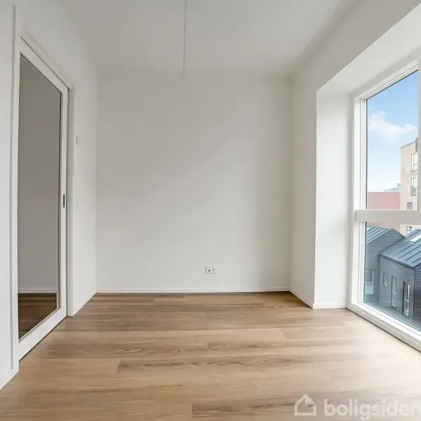 Et tomt rum med trægulv, der reflekteres i et spejl på en indbygget skabslåge. Store vinduer til højre giver udsigt til hustage og himmel.