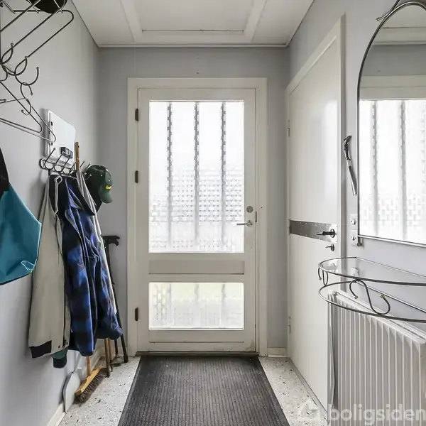 Dørmatte ligger på gulvet foran en hvid dør med sprosser. Jakker og tasker hænger på vægkroge til venstre. Et spejl og en radiator er til højre.