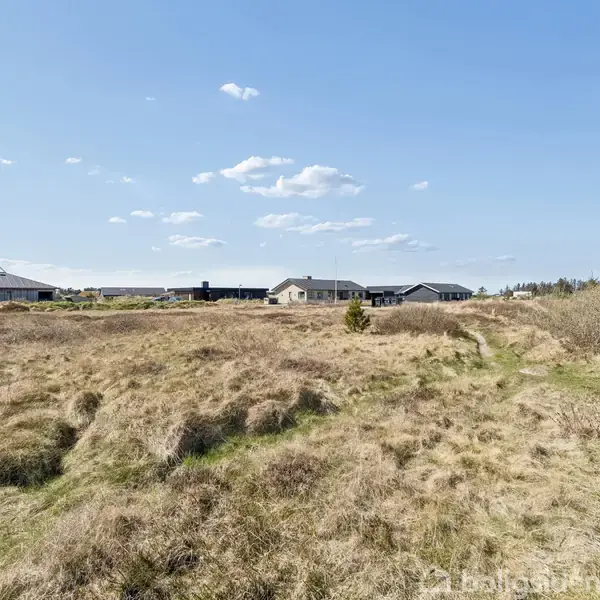 Landskab med åbent, tørt græsareal, der strækker sig mod lavtliggende huse i det fjerne under en klar, blå himmel med få skyer; strukturerne indikerer en landlig eller kystnær beliggenhed.