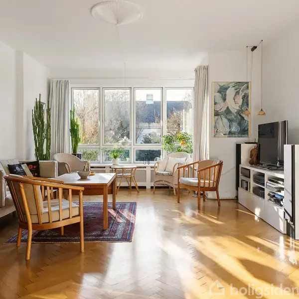 Stue med trægulv og lyse vægge; indeholder sofaer og stole arrangeret omkring et sofabord. Store vinduer tillader naturligt lys, og en reol med tv står til højre.