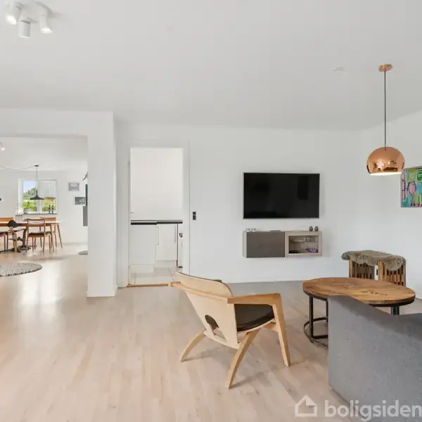 Stue med sofa, lænestol, rundt sofabord. TV på væggen, planter og kunst som dekoration. Lysindfald gennem åbent rum til spisestue med spisebord. Trægulve og lyse vægge.