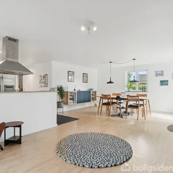 Stol står ved siden af køkkenø, der har en emhætte. Rundt tæppe på trægulv. Spisebord og stole er placeret i et lyst rum med billeder på væggen.