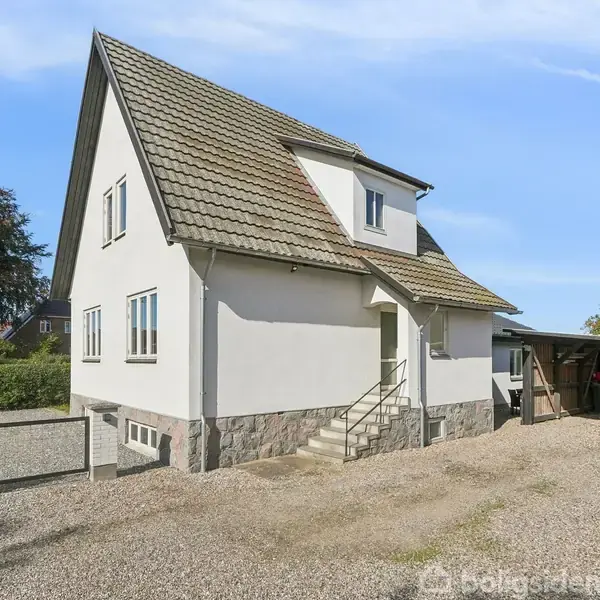 En hvid to-etagers villa med skråt tag står stille i en grusbelagt gårdsplads, omgivet af en hæk og et skur på højre side under en klar blå himmel.