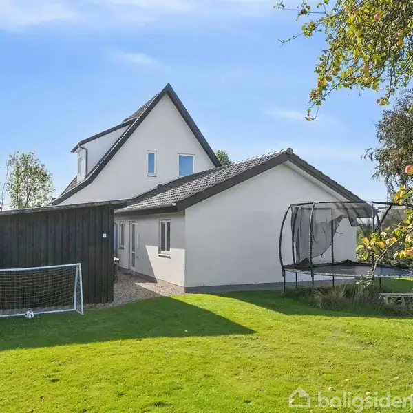 Hus med skråt tag, stående i en have med trampolin og fodboldmål. Sollys skinner over græsplænen, omgivet af træer og buske.