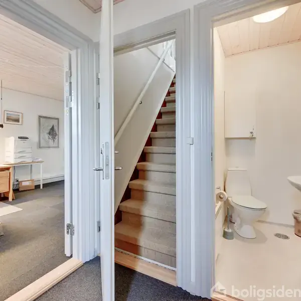 En trappe fører opad mellem to åbne døre; til venstre ses et kontorområde med møbler, og til højre er et lille badeværelse med toilet, vask og brusebad.
