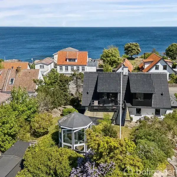 En sortmalet to-etagers hus med skråt tag, har en træbro som fører til hoveddøren. I baggrunden ses en terrasse med havemøbler, omgivet af grønne planter og buske.