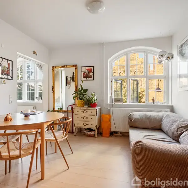 Rundt spisebord med stole står i en stue. Til højre en grå sofa. Store vinduer giver lysindfald. Vægge dekoreret med billeder. En kommode med planter står ved et spejl.