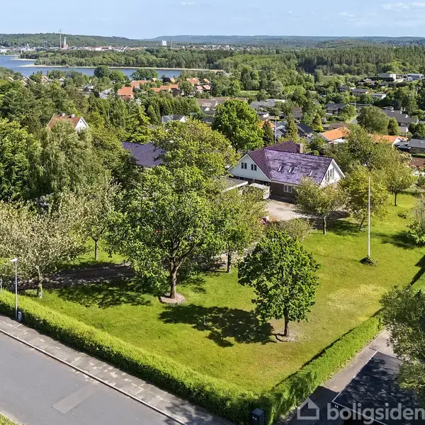 Et hus med en stor have omgivet af træer og buske, placeret i et fredeligt villakvarter med udsigt til en fjord og skovklædte bakker i baggrunden.