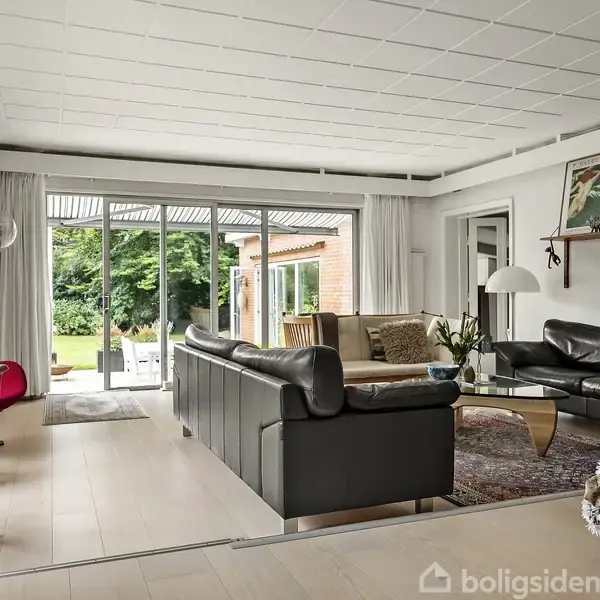 Sort lædersofa står i en lys dagligstue med glasdøre mod en grøn have; omgivet af stole, tæpper og hylder med dekorationer. En rød stol står ved siden af en metalvogn.