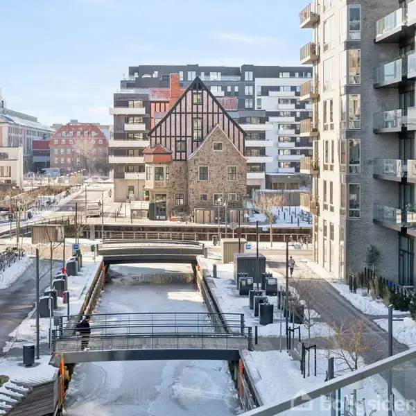 En kanal dækket af sne løber mellem moderne lejlighedsbygninger. Midt i billedet står et gammelt bindingsværkshus. Broer forbinder gaderne på hver side af kanalen.