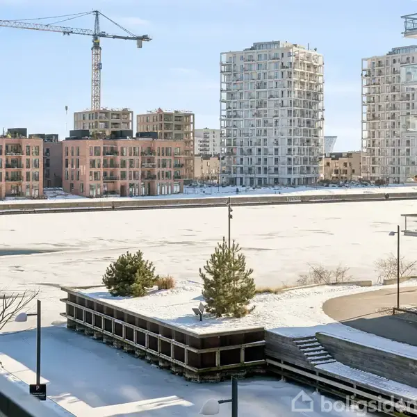 Flod med isdække, omkranset af moderne boligbygninger; en byggekran ses i baggrunden. Sne dækker jorden, og nogle træer står i forgrunden.