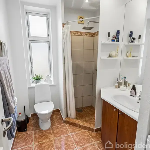 Badeværelse med brusekabine og forhæng til højre, toilet foran vindue til venstre, vaskemaskine og tørretumbler stablet til højre, håndvask med spejl og hylder. Brune fliser på gulvet.