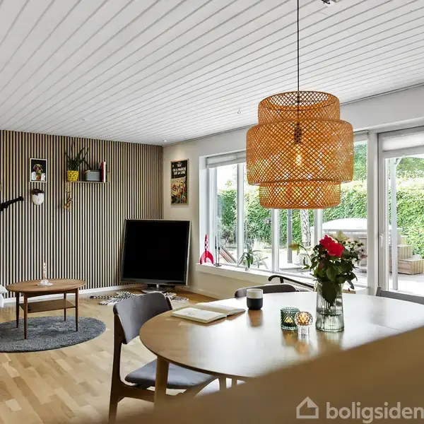 Et moderne stueområde med en rund spisebord, over hvilket der hænger en flettet lampe. Sofasæt og tv står mod en stribet væg. Store vinduer viser grønne omgivelser udenfor.