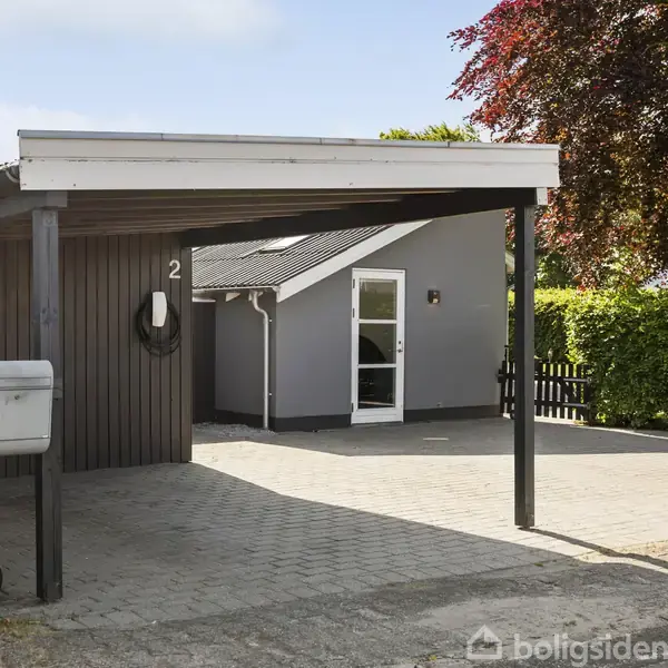En carport med nummer "2" huser en postkasse og beskytter en indkørsel, der fører til et gråt hus med hvid dør. Omkranset af grønne hække og træer.