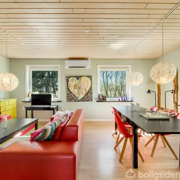 Stue med røde sofaer og et sort spisebord. Væggene har farverig kunst. Store vinduer udsigt til træer. Runde loftslamper lyser rummet. Rummet er lyst med træloft og lyse gulve.