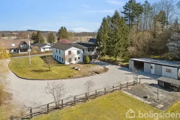 En hvid to-etagers bygning står blandt store træer. En grusvej fører dertil, omgivet af græsplæner, mens en garage står til højre i et landligt område.