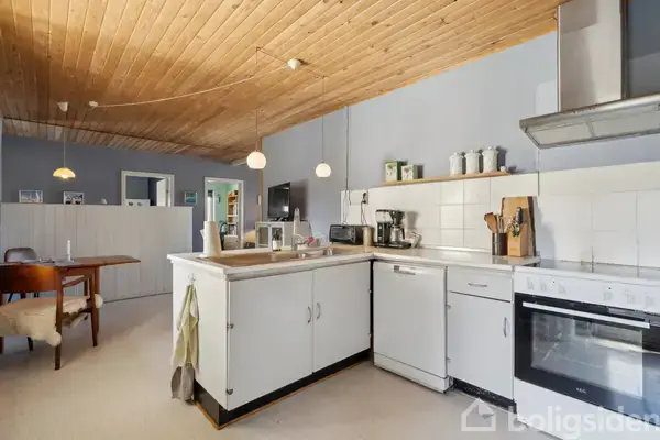 Køkken med hvide skabe og komfur til højre, midt i en træbeklædt loftsrum. Til venstre et spisebord med stole. Hyggelig belysning med hængelamper. Ingen tekst fundet.