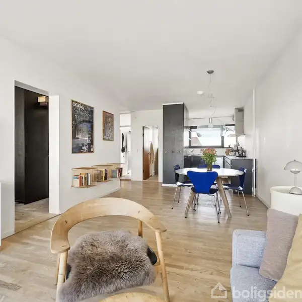 Stue med sofa og lænestol, fladskærms-TV på reol, spisebord med blå stole, og kunst på væggene. Moderne køkken ses i baggrunden, alt i lyse farver.