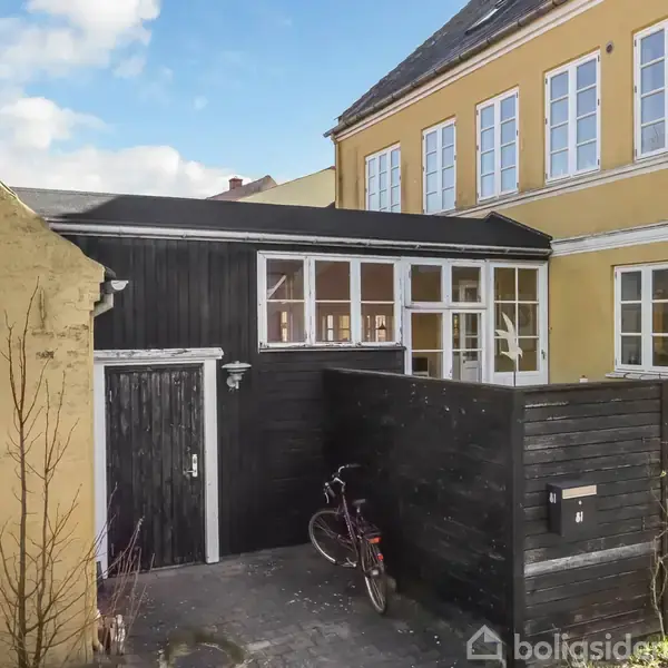 Gult to-etagers hus med flere hvide vinduer. Sort skur med tilstødende dør og hegn. En cykel står parkeret op ad hegnet i en brostensbelagt gård.