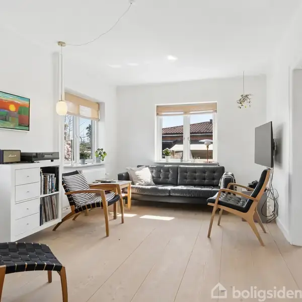 Stue med sort lædersofa og tre stole i moderne design. Væggen har et farverigt maleri. Rummet er lyst med træparketgulv og to vinduer, der lukker naturligt lys ind.