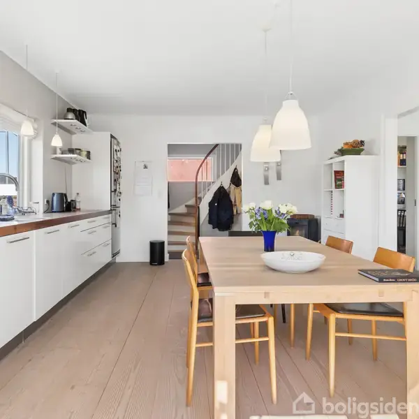 Et lyst køkken-alrum med hvide skabe og træbord. Seks stole står omkring bordet, og der er udsigt til haven gennem store vinduer. Trappen fører til næste etage.