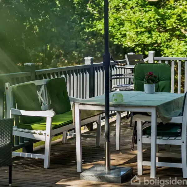Udendørs terrasse med et bord dækket af en dug, omgivet af stole med grønne hynder. I baggrunden ses et sort rækværk og frodig grøn vegetation.
