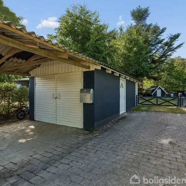 Carport ved siden af en hvid bygning med blå vægge og en skydedør. Omkring bygningen er der flisebelægning og tæt beplantning, samt et plankeværk i baggrunden.