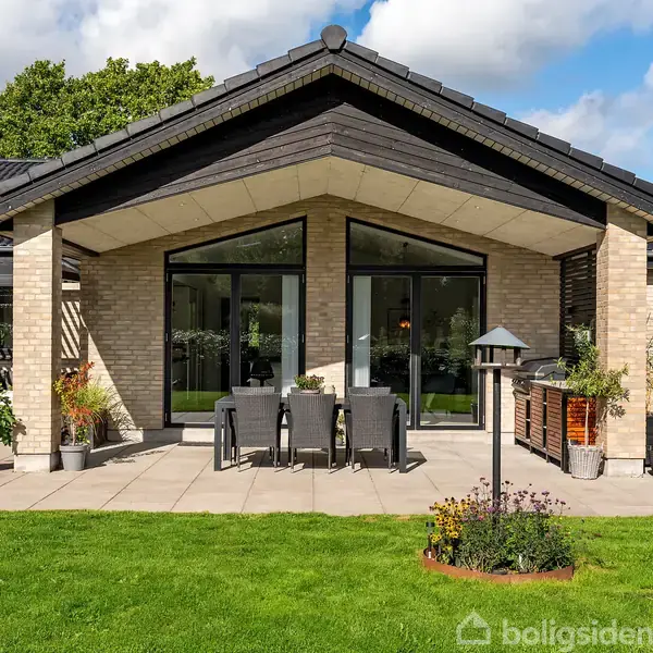 En moderne villa med store glasdøre åbner ud til en flisebelagt terrasse. Havemøbler og planter dekorerer området, der ligger i en grøn have under en blå himmel.