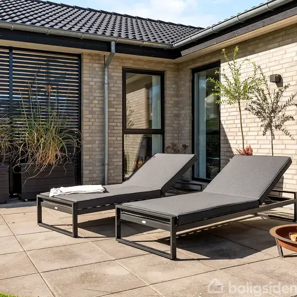 To solsenge står på en flisebelagt terrasse ved et moderne murstenshus. En ildskål er placeret ved siden af, og der er høje planter langs husvæggen.