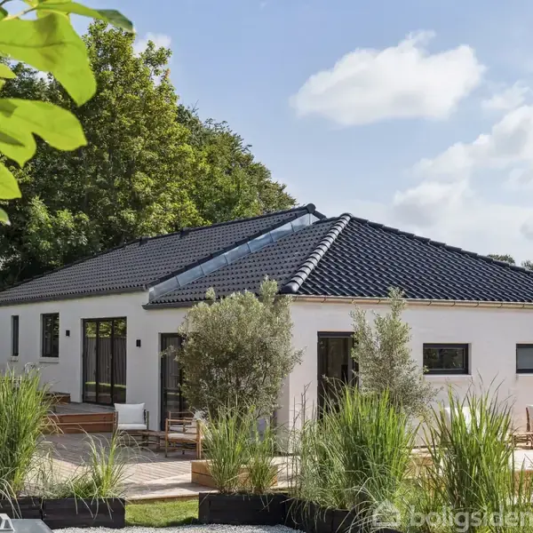Et hvidt hus med sort tag står i en have med grønne planter og terrasseplanker. Flere store vinduer åbner til en solrig dag med en let overskyet himmel.